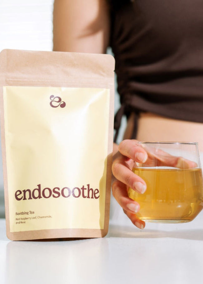 A person holds a glass of amber-colored Endosoothe tea next to its kraft paper stand-up pouch packaging on a white surface.