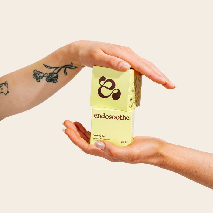 Two hands holding a small pale yellow Endosoothe Soothing Cream box against a solid light beige background.